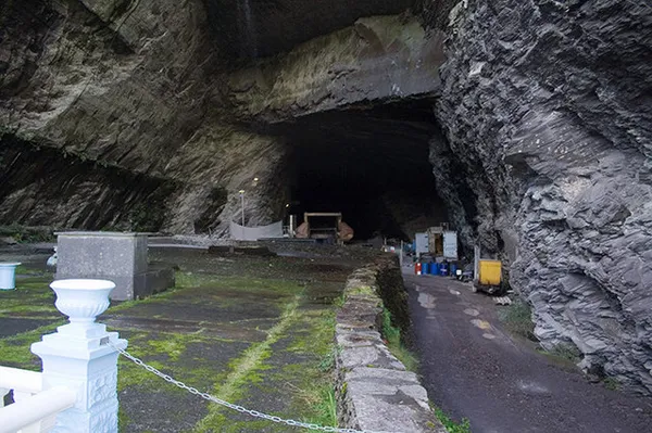 Une grande ouverture sombre ressemblant à une grotte dans une falaise d'ardoise, avec un chemin pavé et une balustrade blanche devant.