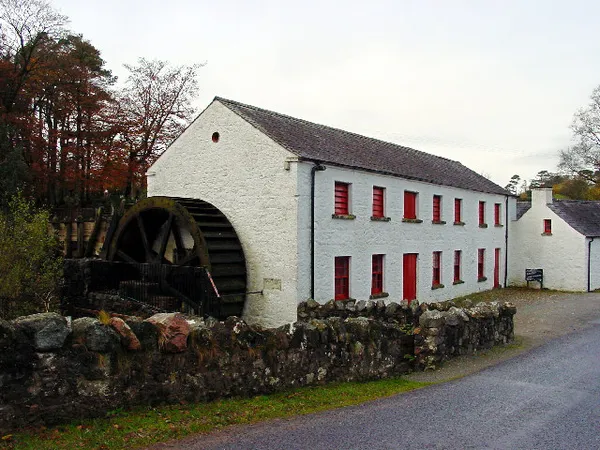 Eine lange weiß getünchte Mühle mit roten Fensterrahmen und einem großen hölzernen Wasserrad hinter einer Steinmauer.