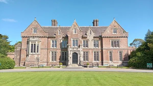 A large red brick Victorian house with gables and tall chimneys behind a mown lawn under blue sky.