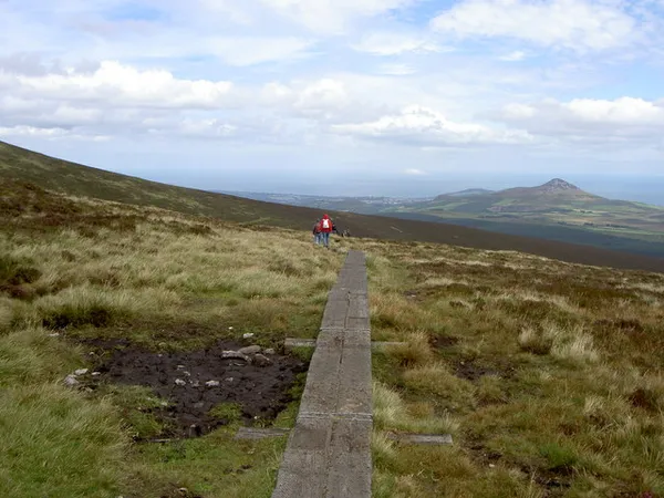 A narrow wooden boardwalk crossing open heather and bog towards a distant walker and a pointed hill.