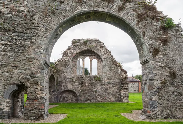 Un arco de piedra en ruinas que enmarca un hastial sin techo con una ventana de tres luces, situado en un césped verde.